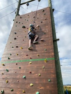 Picture shows a young girl wearing splints scaling an outdoor wooden climbing wall while attached to a harness.